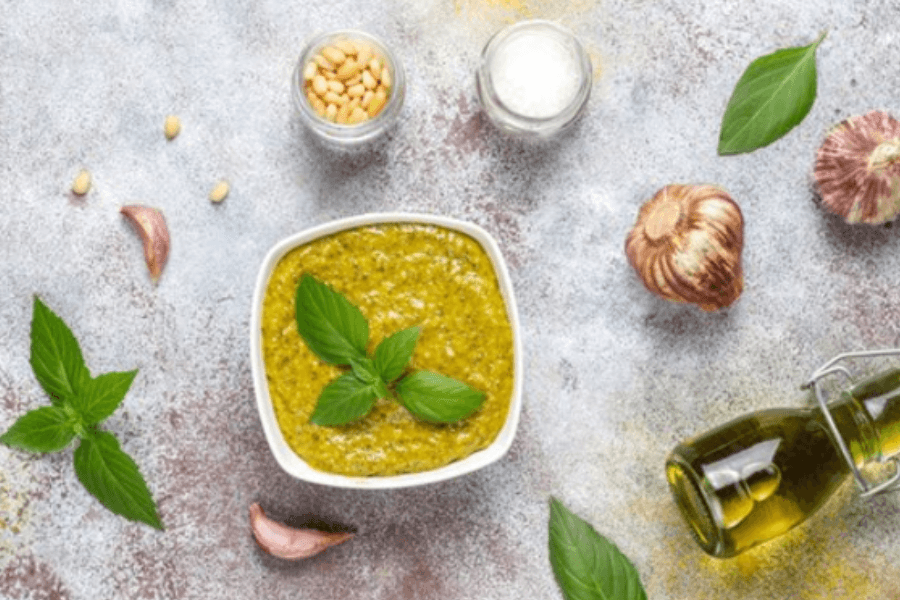 A bowl of fresh basil pesto garnished with basil leaves, surrounded by its key ingredients—pine nuts, grated Parmesan, garlic cloves, olive oil, and fresh basil leaves—arranged artistically on a textured surface.
