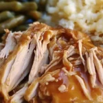 A plate with shredded Mississippi Chicken, mashed potatoes with gravy, and green beans, served as a comforting meal.